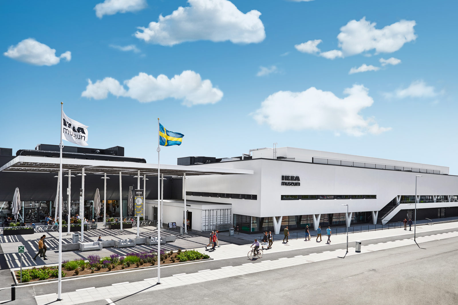 The white IKEA Museum building under a blue sky, people walking, one IKEA flag and one Swedish flag on a windy, sunny day.