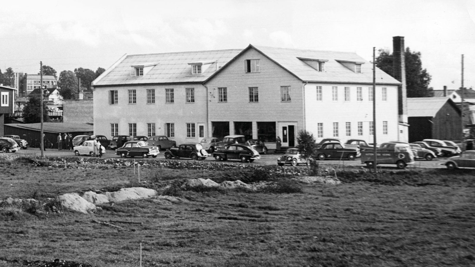 Stor sliten grå byggnad med parkeringsplats, 1940-talsbilar.