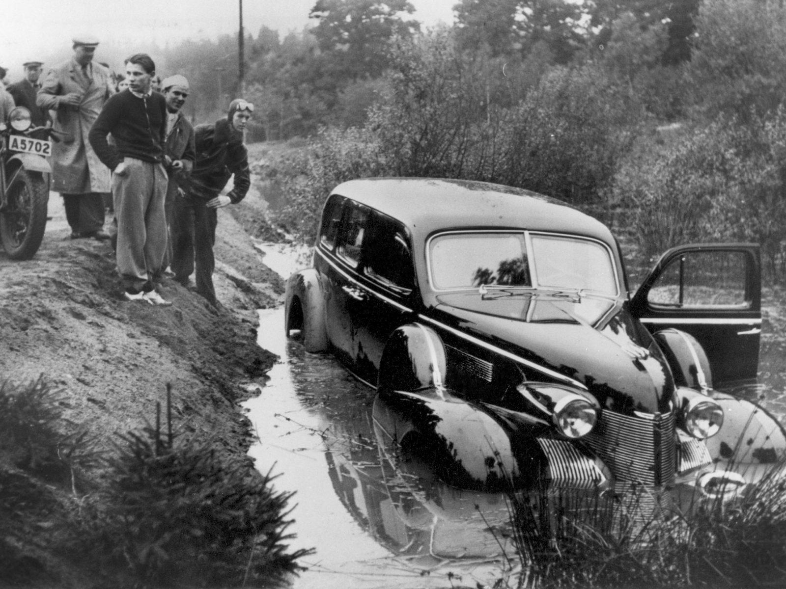 A 1940s Cadillac sits in water-filled roadside ditch, male onlookers in 1940s clothing have gathered on the road above.