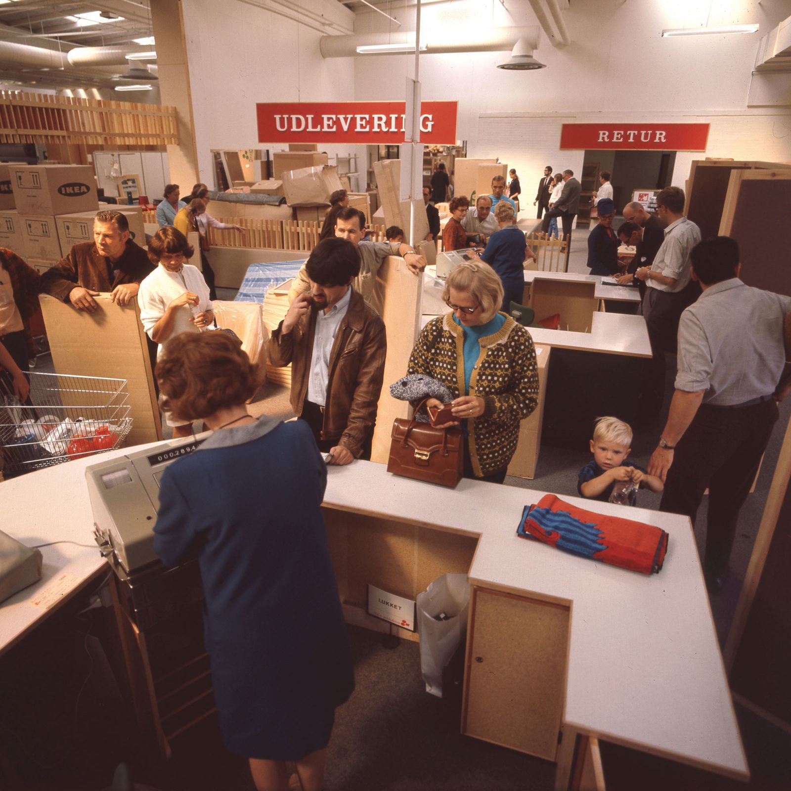 Busy, disorganised checkout area, 1960s IKEA store. In background, big signs in Norwegian read Delivery and Returns.