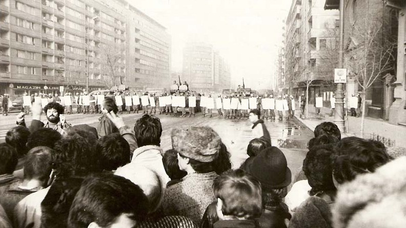 Manifestants dans une large rue, lèvent le poing contre une ligne serrée de policiers anti-émeute.
