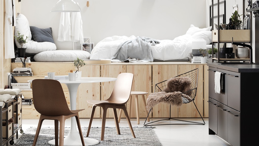 Steps lead up to raised platform in blond wood with bed and seat cushions. Tables and chairs in front, next to kitchen area.