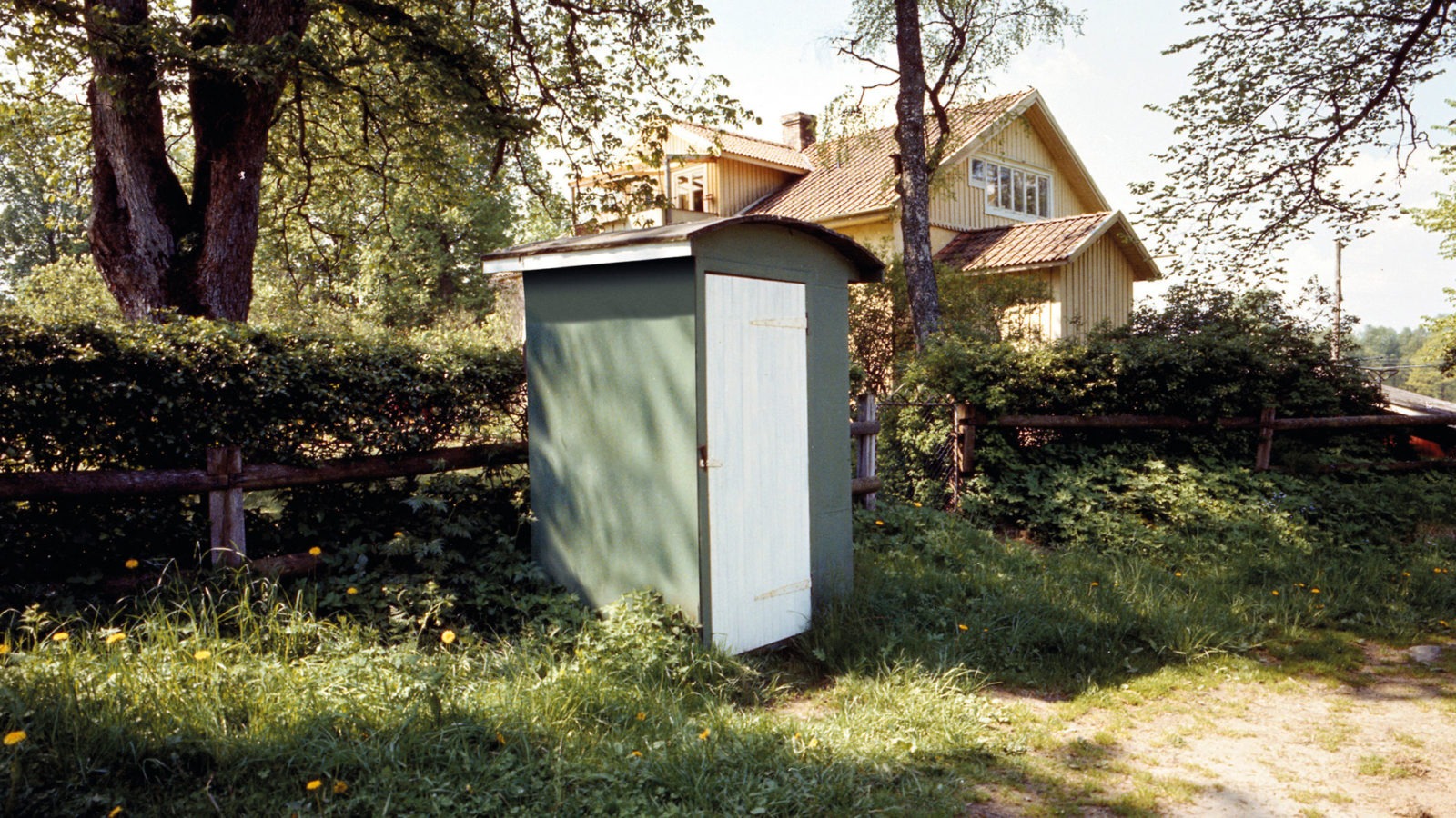 Sonniger Tag, ländliche Gegend, Wiese mit einem Holzzaun und einem grünen Schuppen, im Hintergrund ein großes gelbes Haus.