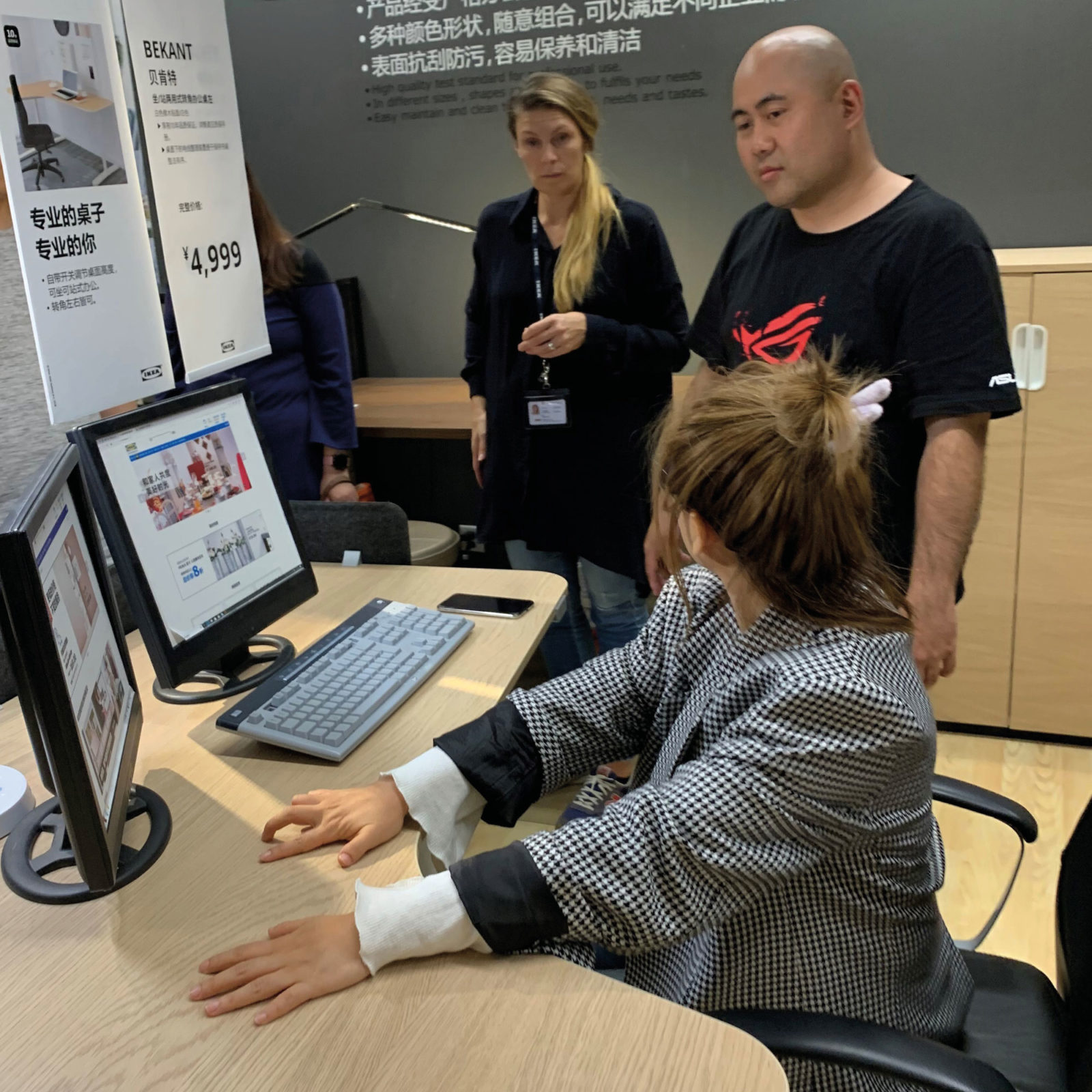 A young dark-haired woman sits at computer and looks up at young man and blonde woman standing next to her.