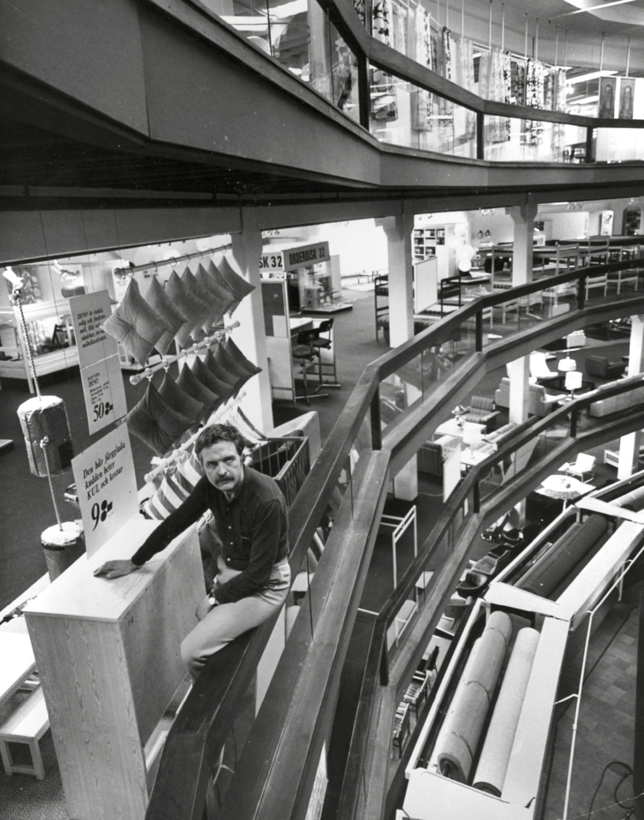 A moustached Hans Ax, casually dressed, seen in store environment, furniture, pillow displays and signs seen in background.