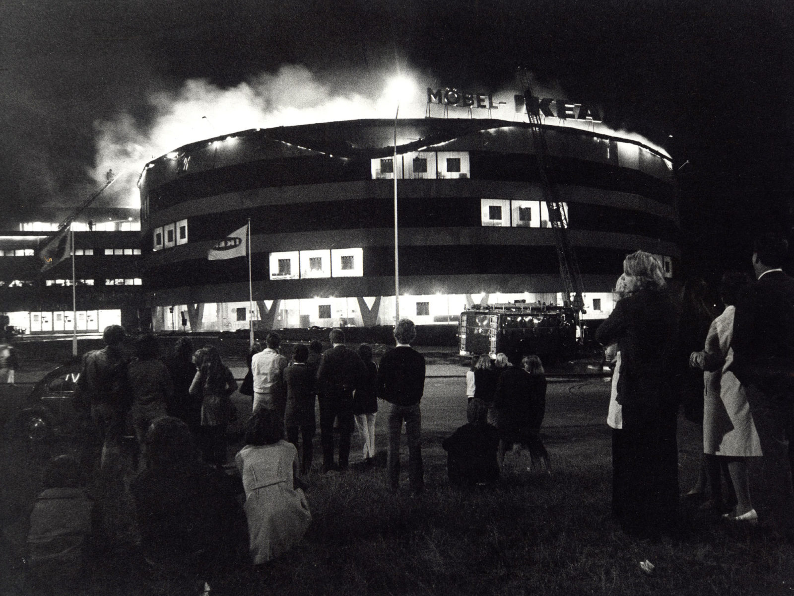 Onlookers gathered at night in front of huge burning building with sign FURNITURE IKEA in Swedish and a fire truck.