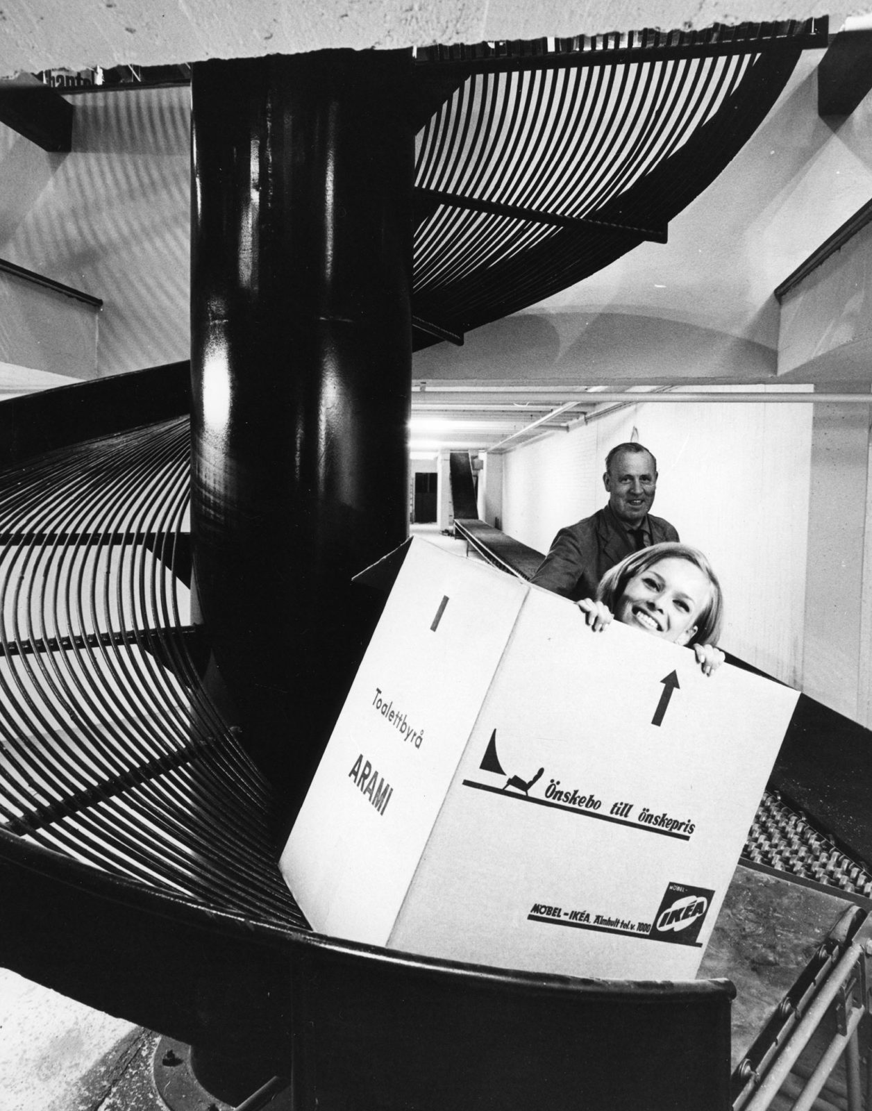 Smiling girl peers out of moving box marked IKEA, sliding down a corkscrew shaped chute towards a conveyor belt.