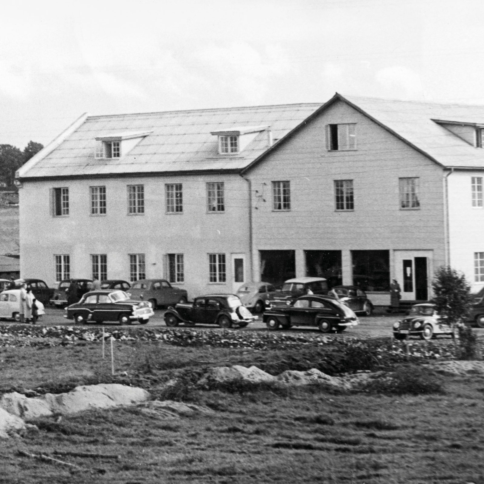 Svartvitt foto av stor vit byggnad, bilar från 1940- och 1950-talet parkerade utanför.