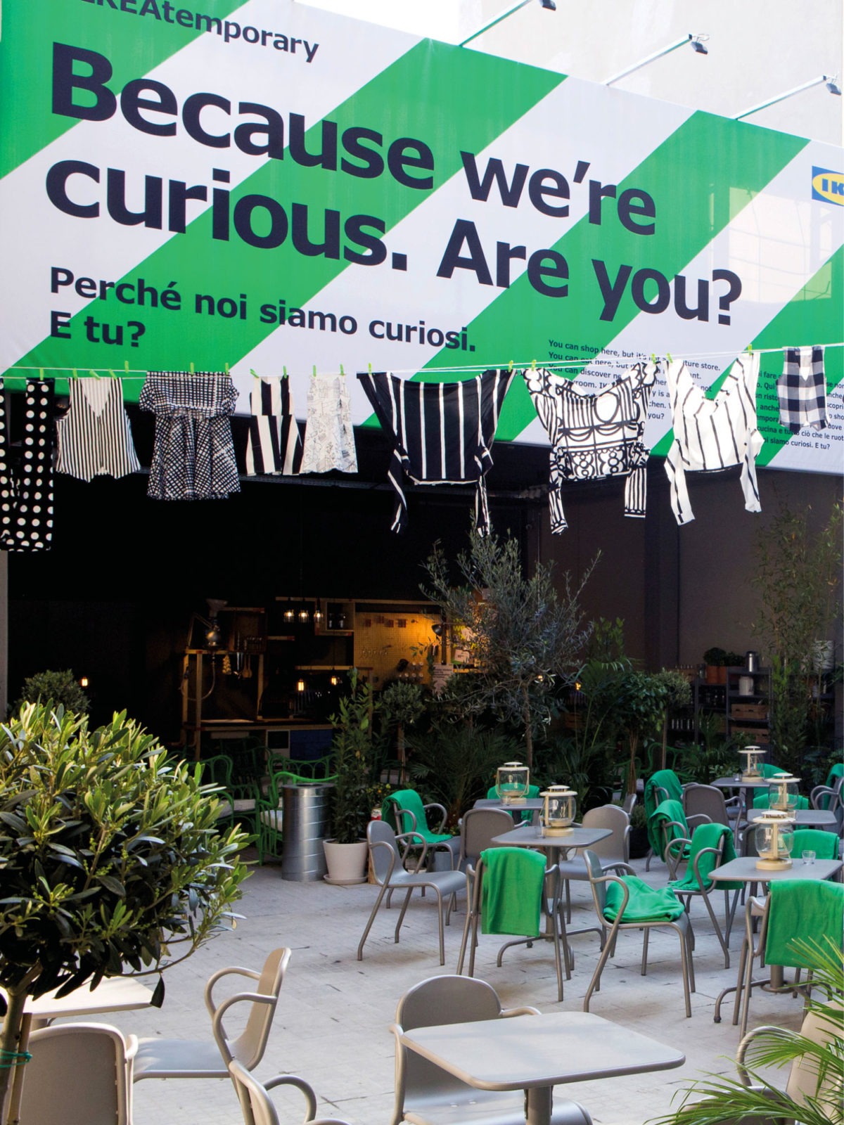 Garments on clothes line hanging over outdoor seating space, in front of big sign saying: Because we are curious. Are you?