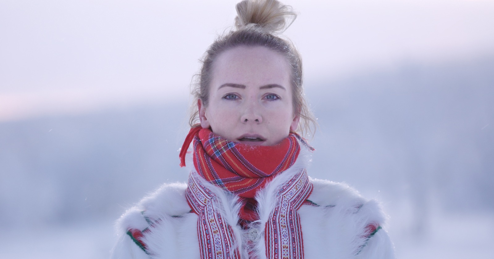Eine junge Frau in einer verschneiten Winterlandschaft, die einen traditionellen lappländischen Mantel aus Rentierfell und einen roten Schal trägt.