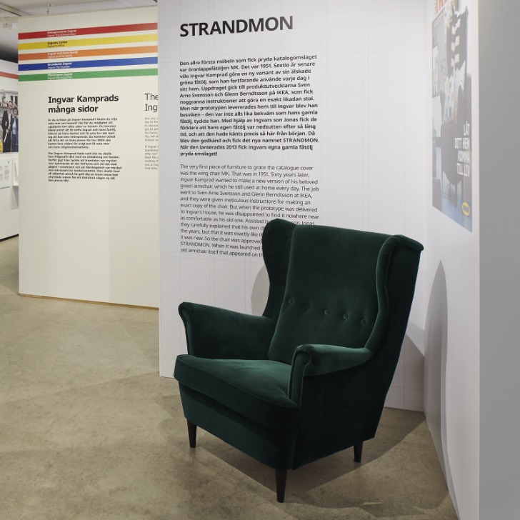 Ein dunkelgrüner STRANDMON Ohrensessel im IKEA Museum. Auf einer großen Tafel dahinter wird die STRANDMON Geschichte erzählt.
