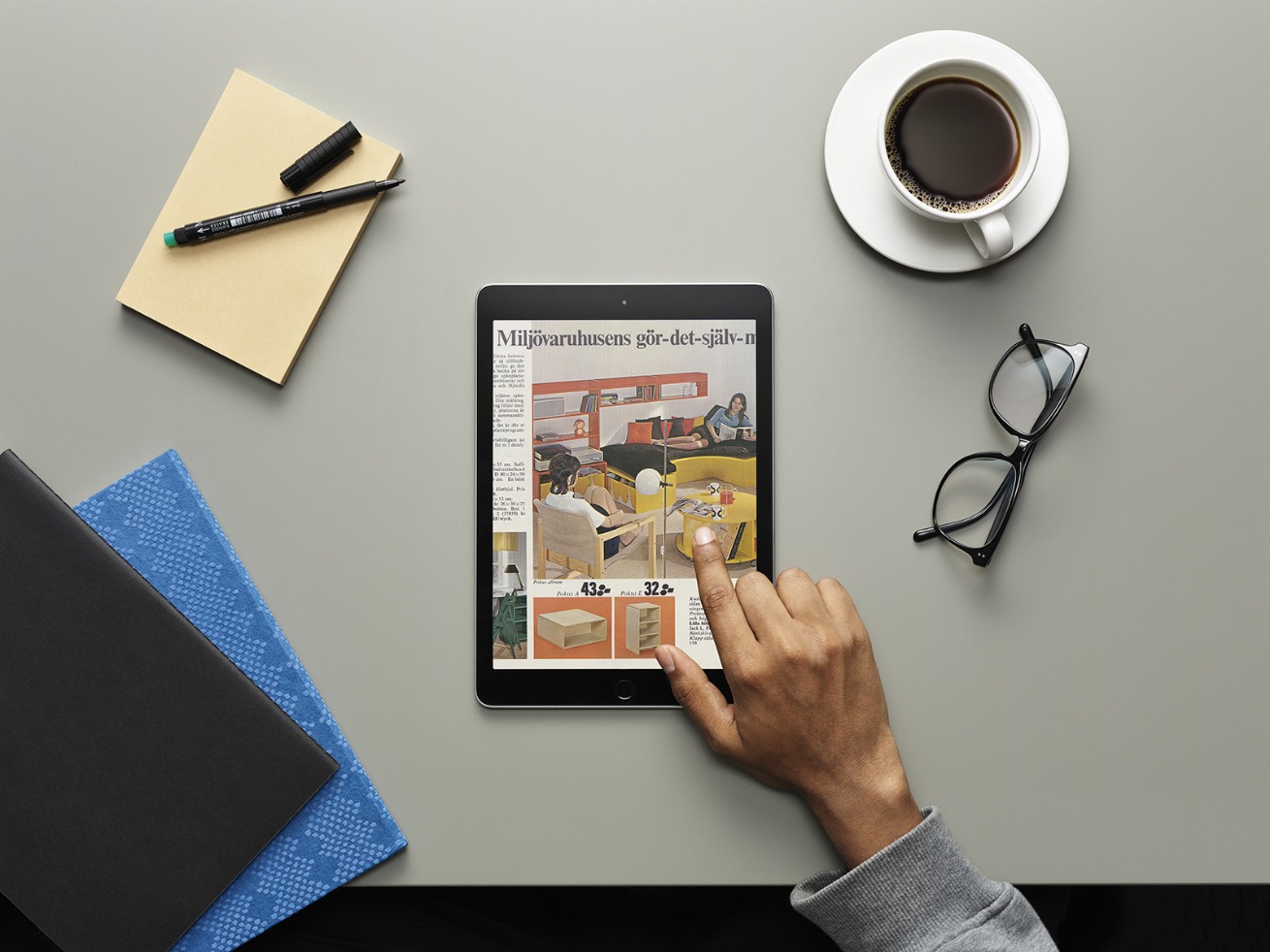 Eine graue Tischplatte, eine Tasse Kaffee, eine Brille, Notizbücher, ein Stift und eine Hand, die mit einem Tablet einen alten IKEA Katalog durchblättert.