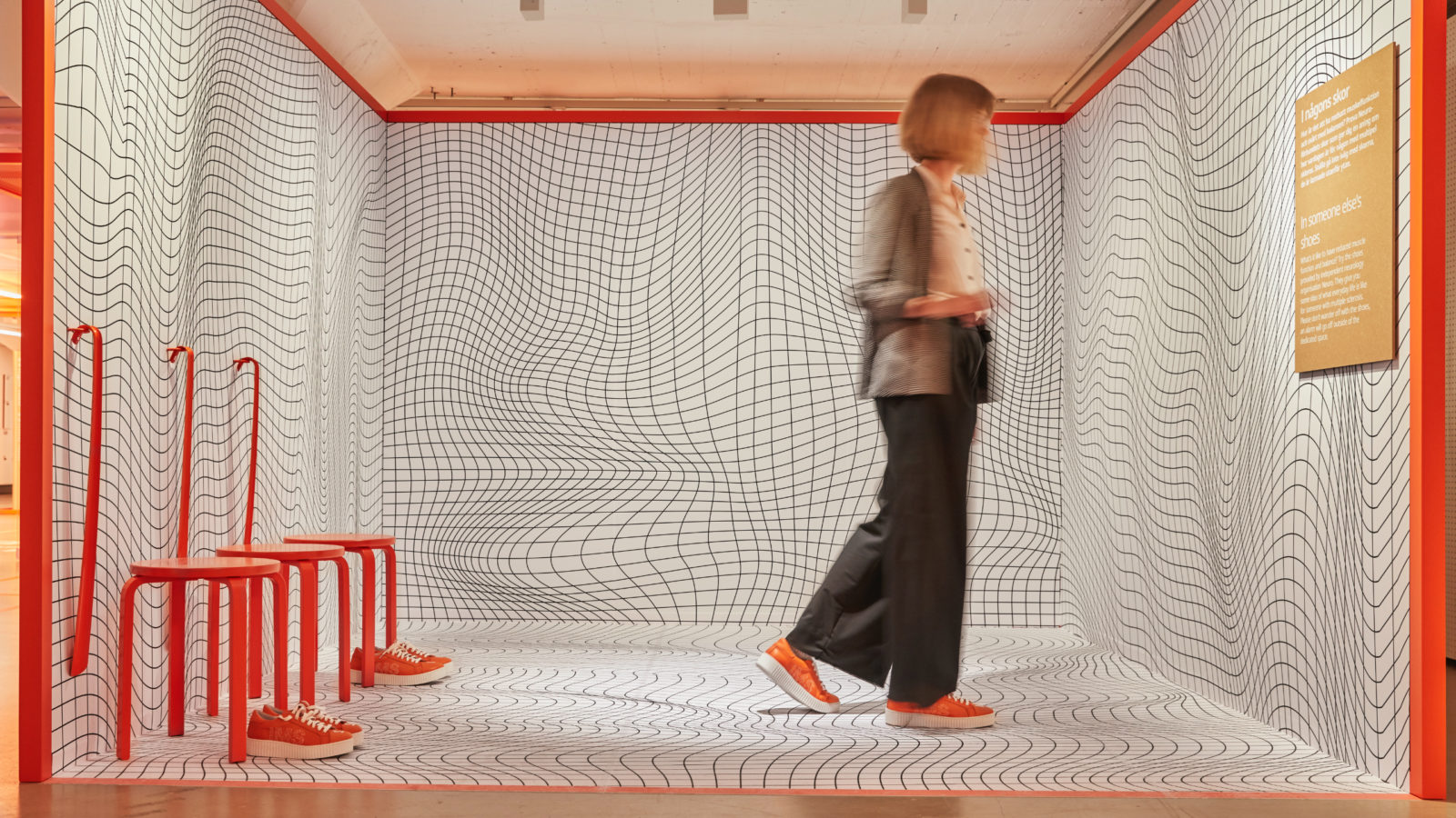 Eine Frau in einem mit schwarz-weißen geometrischen Mustern bemalten Raum, in dem sich jeweils drei leuchtend orangefarbene Hocker, Schuhe und Schuhanzieher befinden.