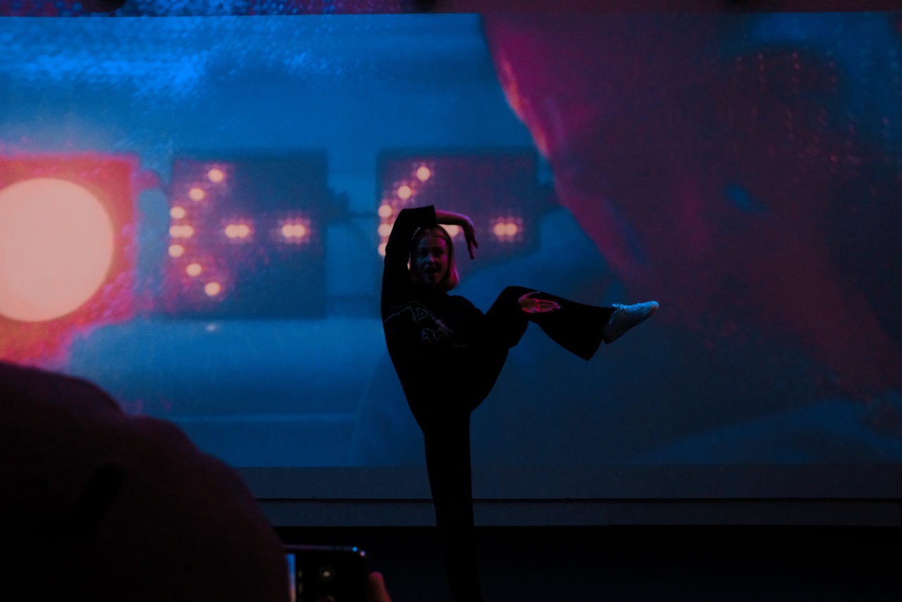 A woman lifting up her leg while dancing in a large, dimly lit room in front of a blue background with arrow-shaped lights.