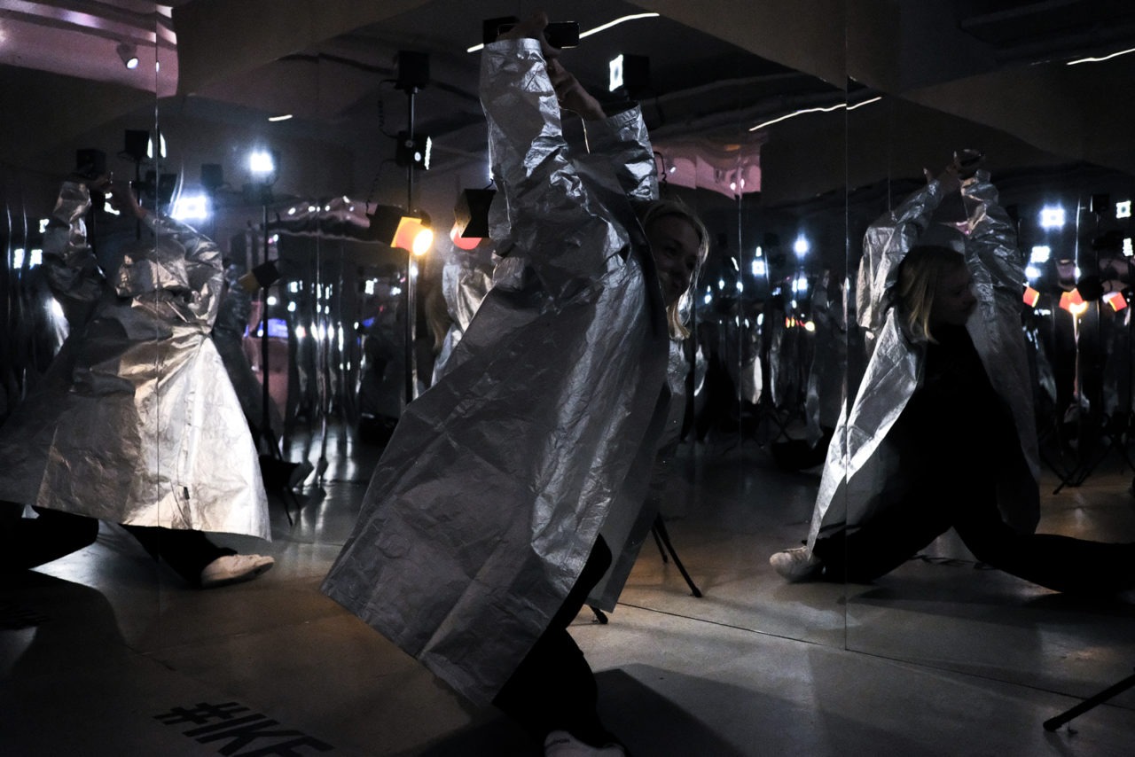 A woman in a long, silvery coat dancing in a room with mirror walls and products from the IKEA FREKVENS collection.