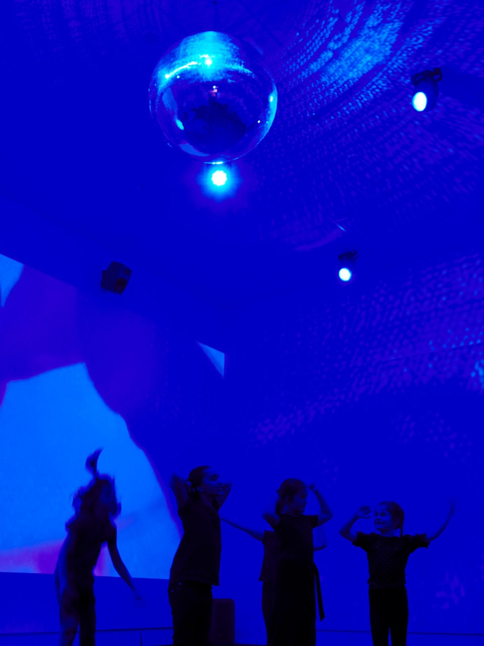 People of different ages dancing in a large room with a blue light and a disco ball hanging from the ceiling.