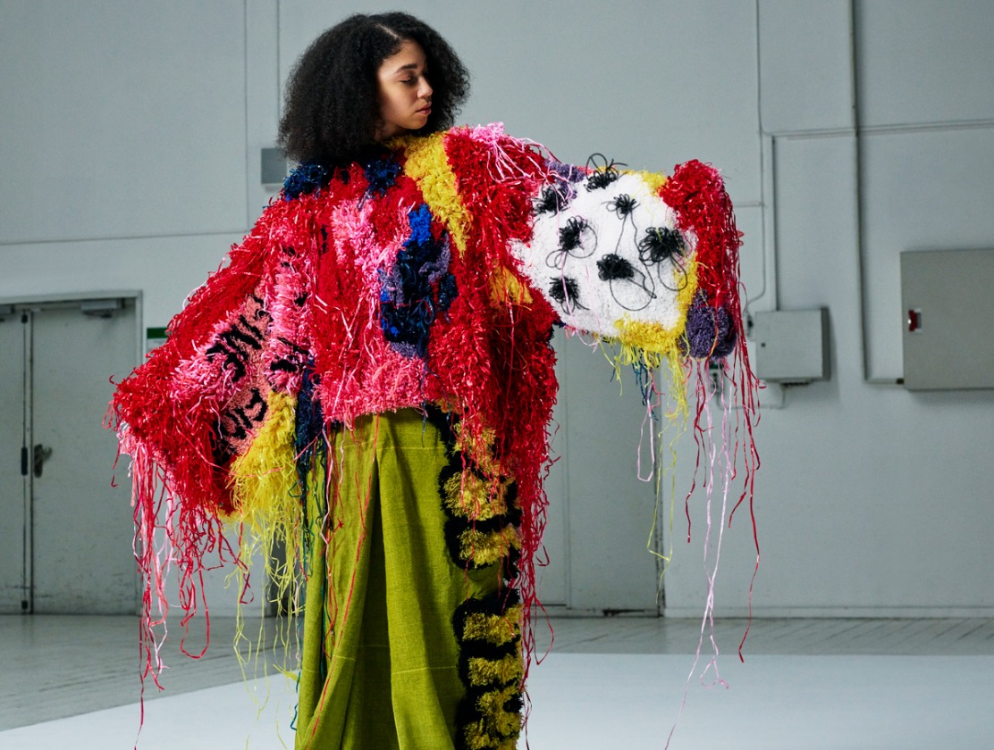 A woman wearing a multicoloured, tufted outfit with long, messy threads and graffiti decorations.