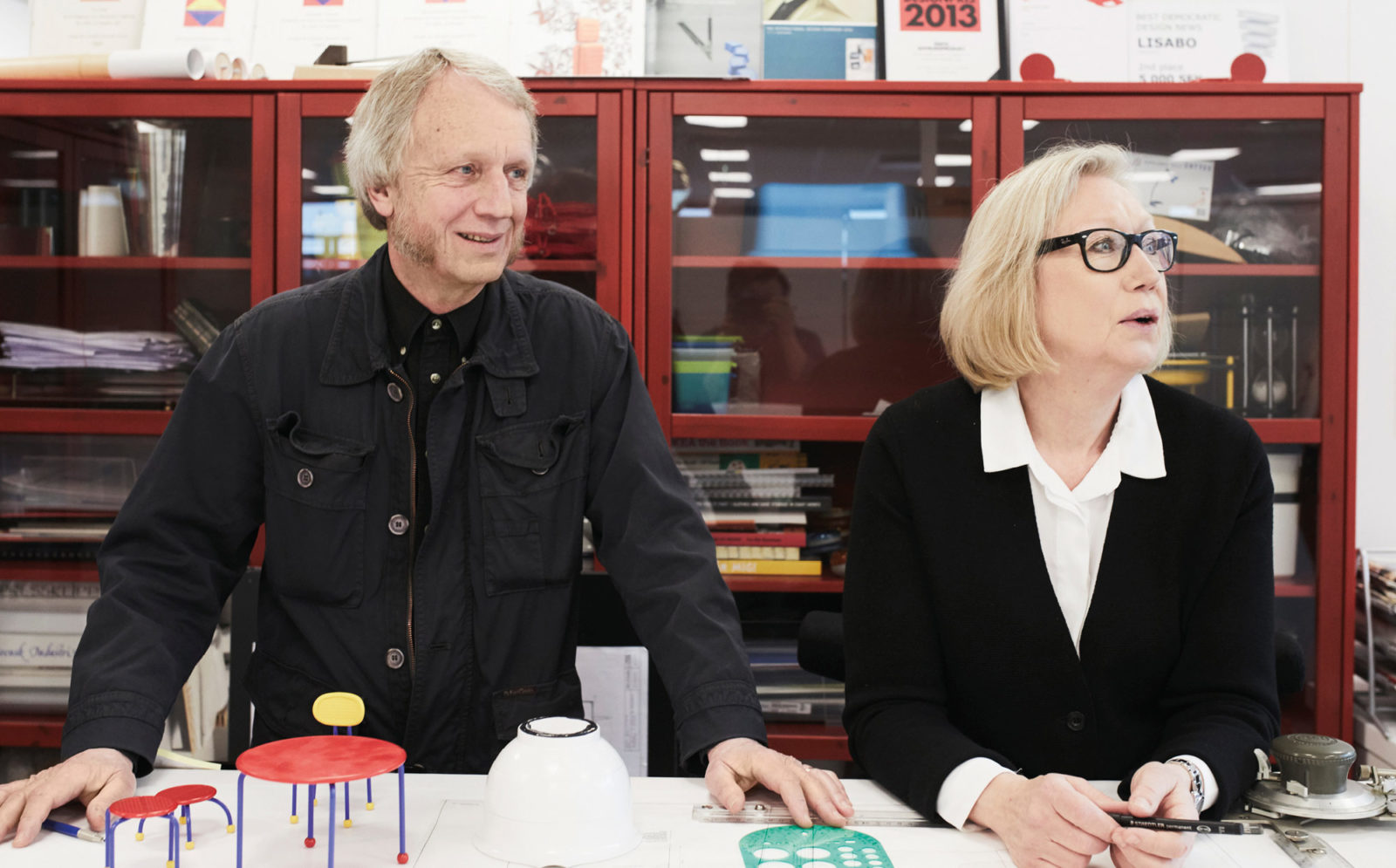 Medelålders man och kvinna med blonda, grånande hår och svarta kläder, står vid ett ritbord med prototyper och linjaler.