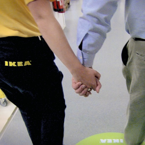 Gros plan sur deux personnes de dos se tenant par la main, une en uniforme, logo IKEA dans le dos, l’autre en chemise bleue.