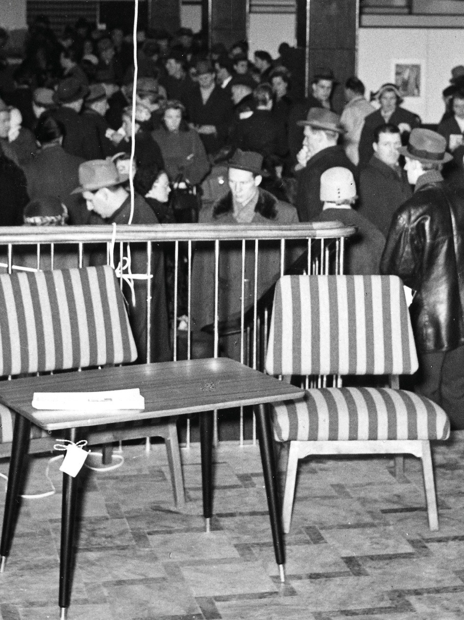 Randig soffa, träbord i 1950-talsstil uppställt i mässlokal. I bakgrunden syns myller av folk.