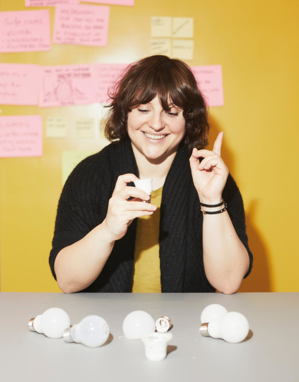 パウリーナ・パヤックが、LED電球のパーツが散らばるテーブルでパーツを1つ手に取り、誇らしげな笑顔で指を１本立てている。