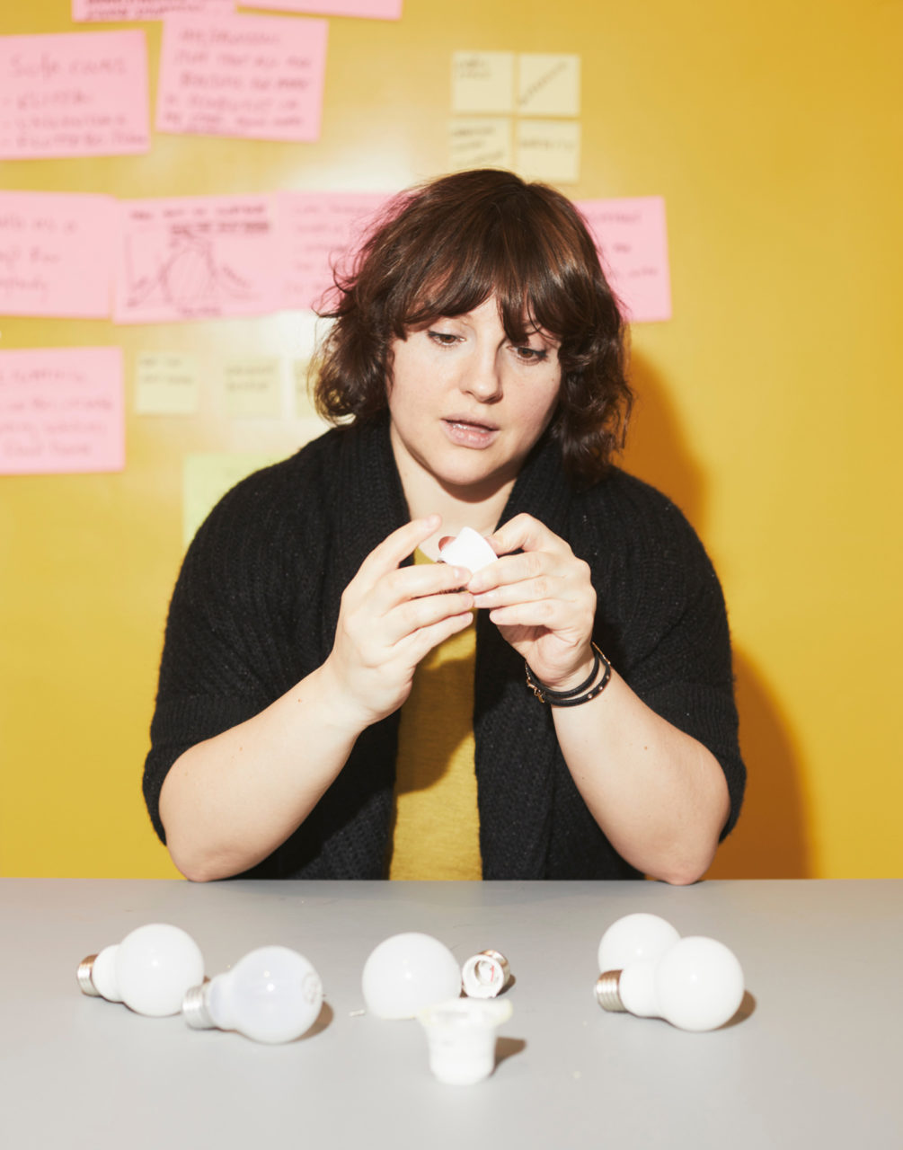 パウリーナ・パヤックがLED電球のパーツが散らばるグレーのテーブルの横に座り、パーツを１つ手に取ってじっくりと観察している。