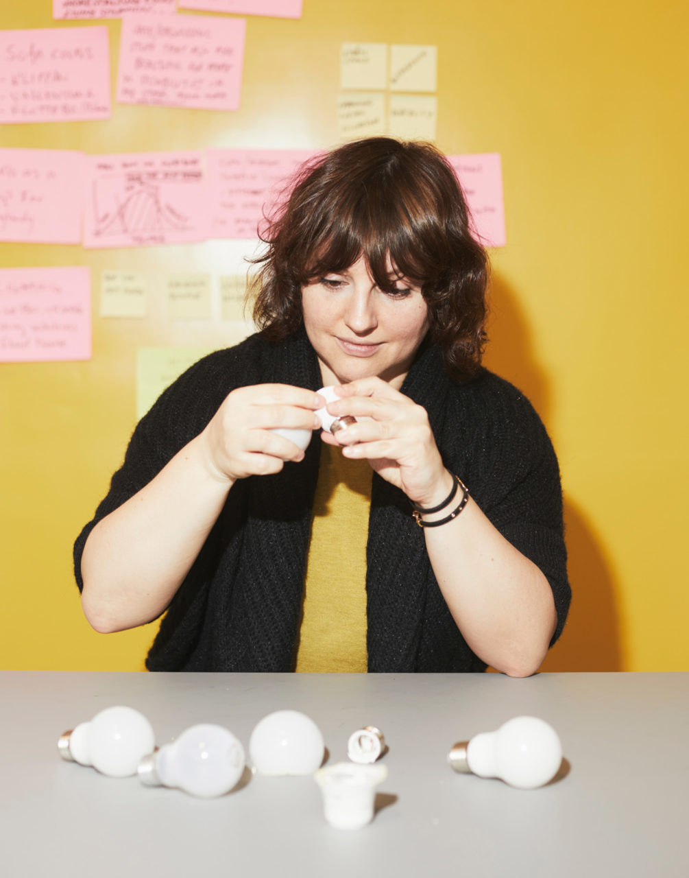 パウリーナ・パヤックがLED電球のパーツが散らばるグレーのテーブルの横に座り、パーツを１つ手に取ってじっくりと観察している。