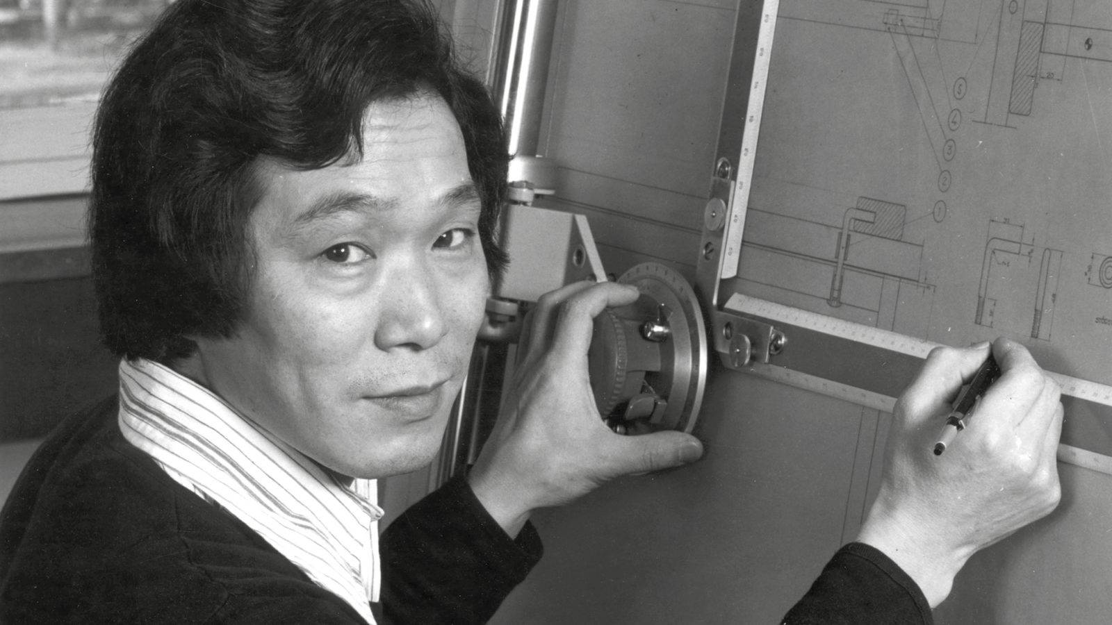 Man with black hair, striped shirt and sweater, working at drawing board.