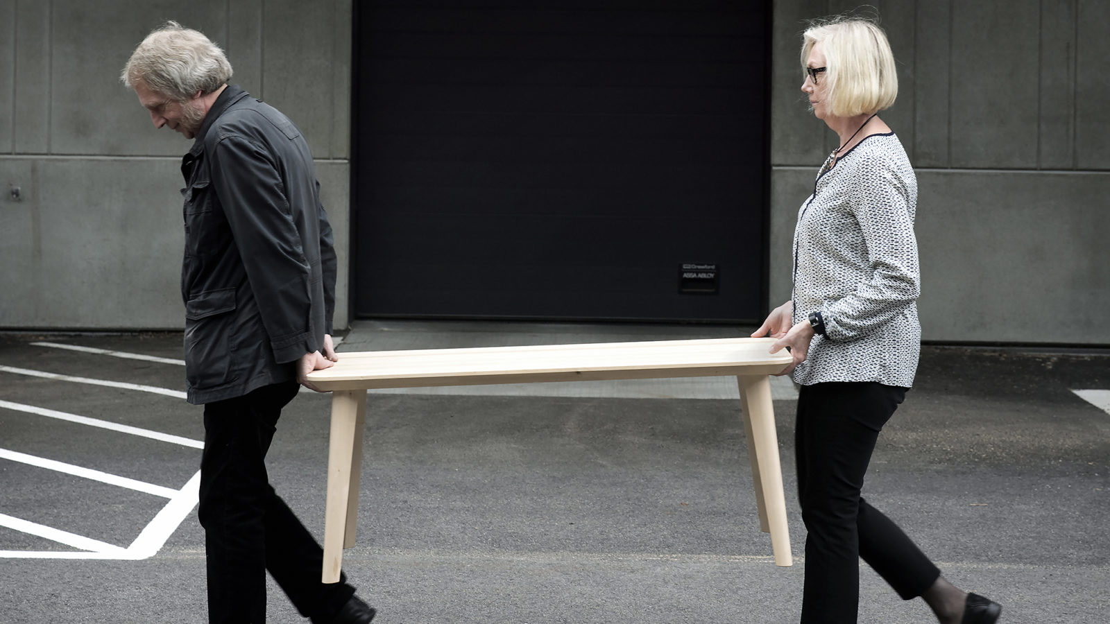 Ageing man and woman dressed in black and white, Knut Hagberg and Marianne Hagberg, carrying simple wooden table together.