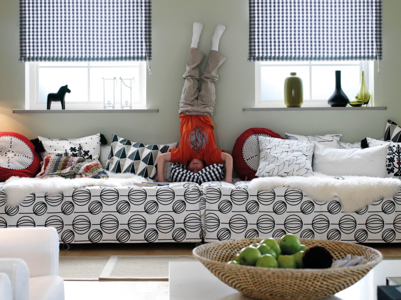 A child is standing on its head right where two end-to-end beds meet. Light textiles in check or graphic patterns.