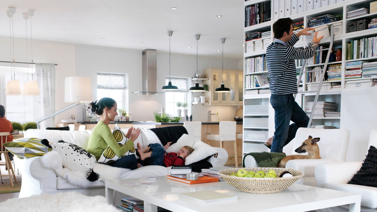 A family with children and a dog spread around a large, light family room with an open-plan solution. The furniture is either light wood or white.