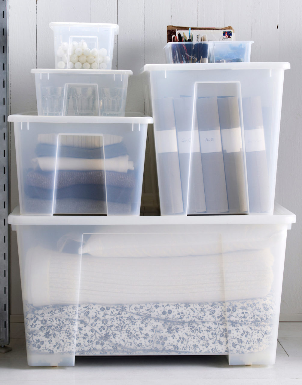 Boîtes en plastique de différentes tailles empilées. Linge de lit, vêtements, dossiers et bougies y sont rangés avec ordre.