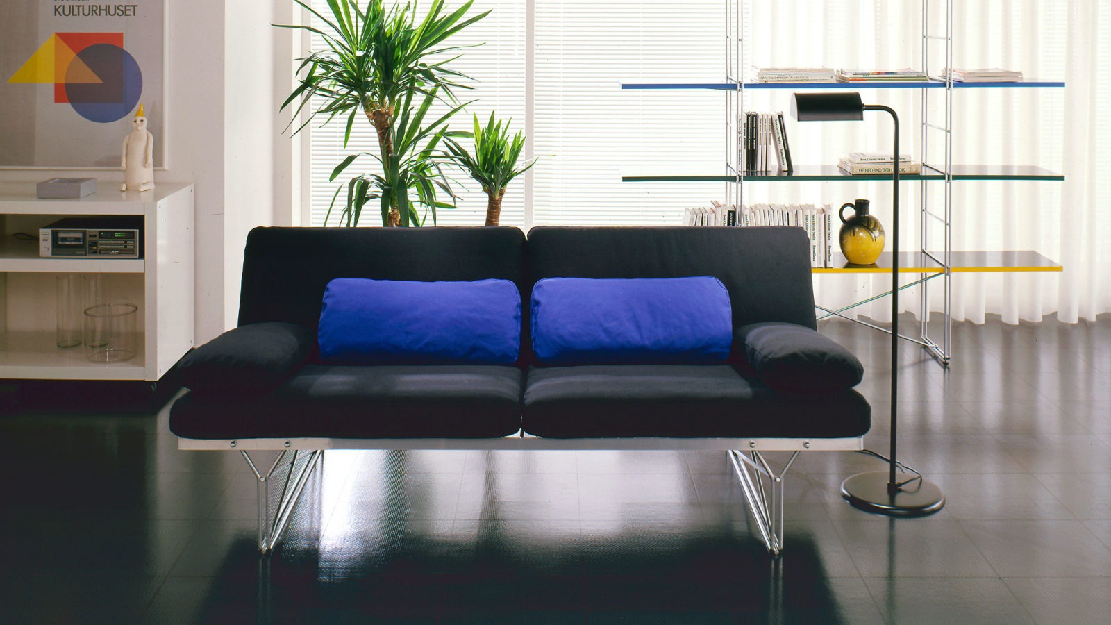 Schwarzes MOMENT Sofa und buntes MOMENT Regal mit dünnen Metallrahmen in einem Raum mit glänzendem, schwarzem Fußboden.