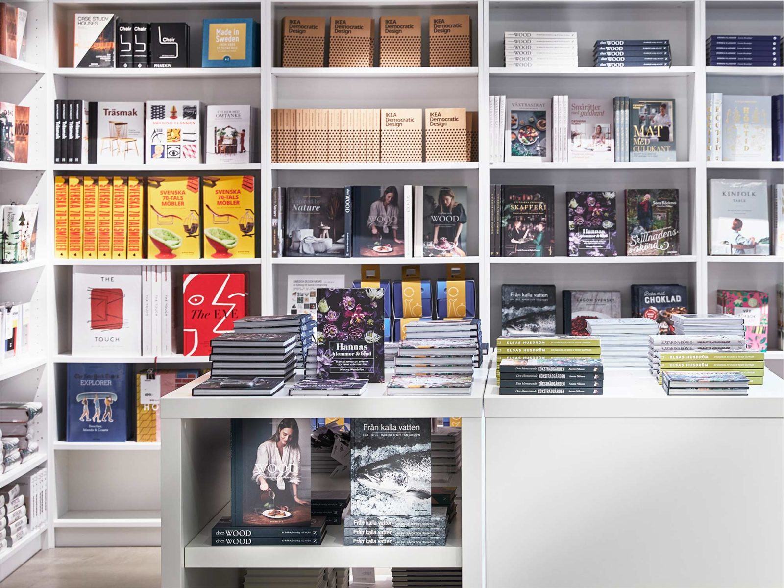 Book section in shop with white shelves, in foreground books are stacked on top of table and on shelves beneath.