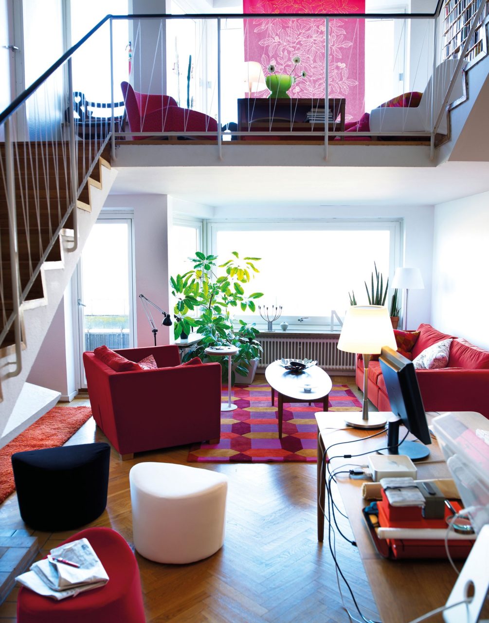 Färgglatt rum i två etage med sittpuffar, sittgrupper i vitt och rött, med gröna växter i fönstren.