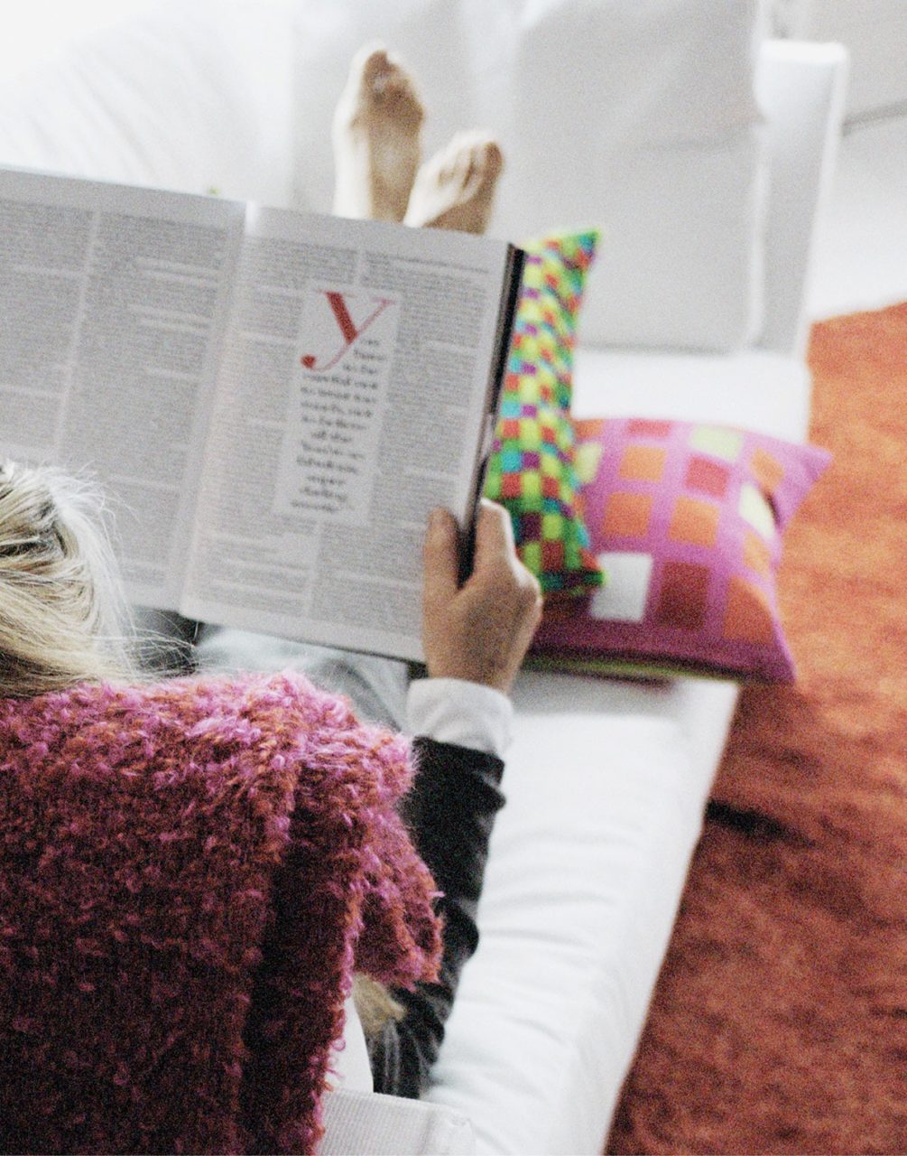 Detaljbild bakifrån av person på en vit soffa som läser tidning och har satt upp sina bara fötter på färgglada kuddar.