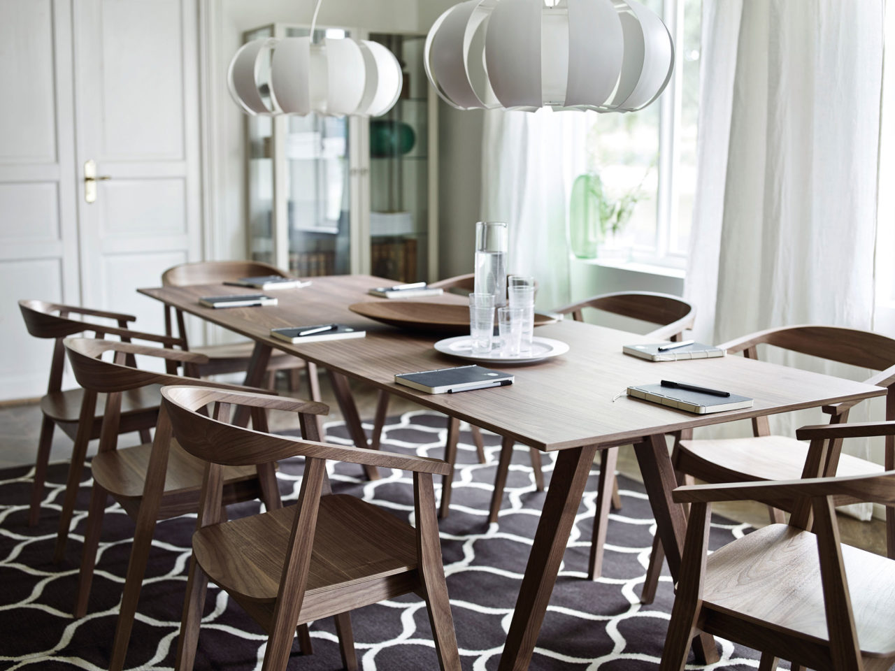 Bruna trästolar runt ett avlångt mötesbord i samma material. Lågt hängande lampor, svarta anteckningsblock vid varje plats.