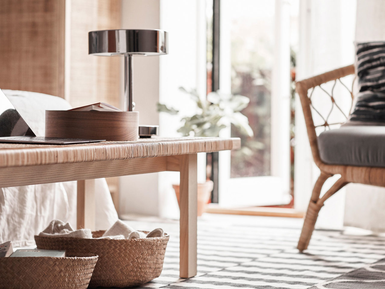Detaljbild av rottingfåtölj, bord i ljust trä och rotting, silverfärgad bordslampa och flätade korgar.