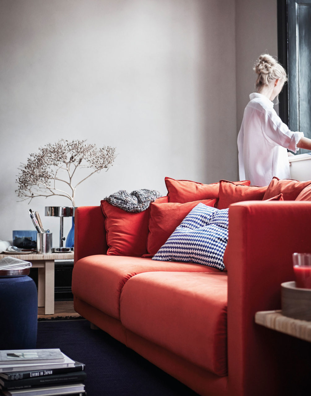 Organgeröd soffa, blomarrangemang i vas, ung kvinna står vid ett fönster.