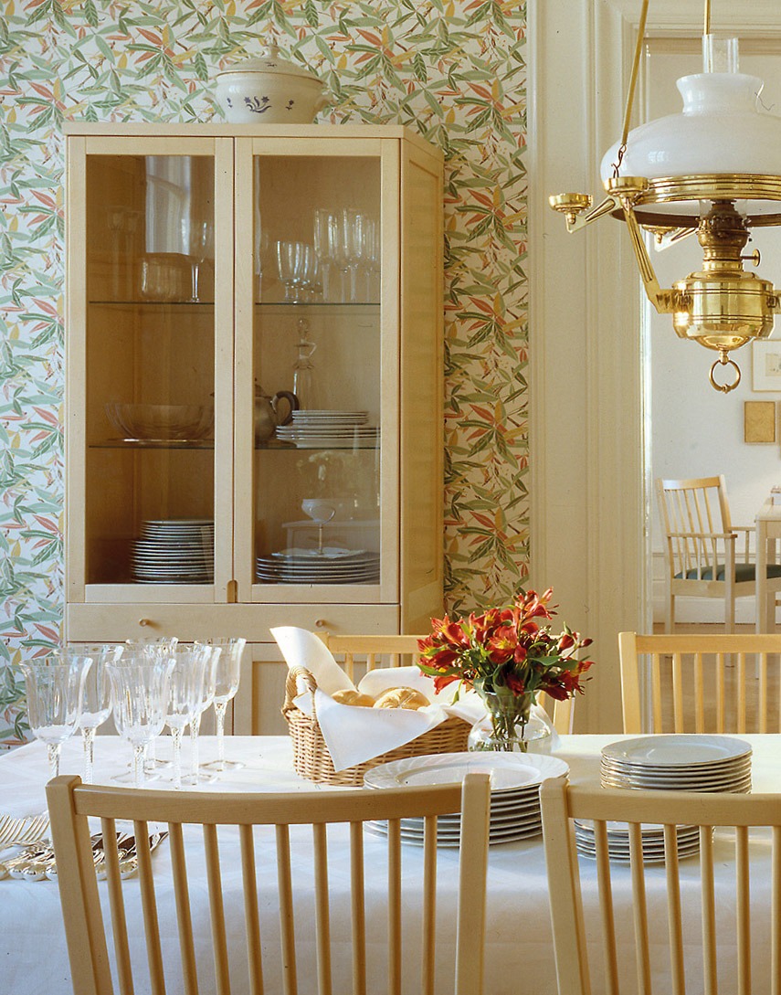 Matrum i vita och beiga toner, vinglas och tallrikar på vitdukat bord, ett vitrinskåp med servis i bakgrunden.