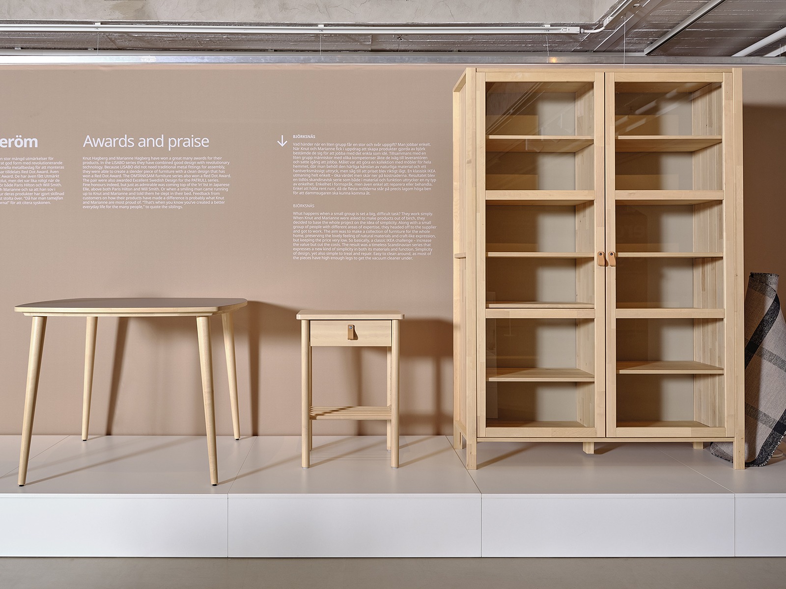 Two desks and a chair in blond wood displayed on a low podium in front of a beige wall with a big close-up image of a wedge dowel.
