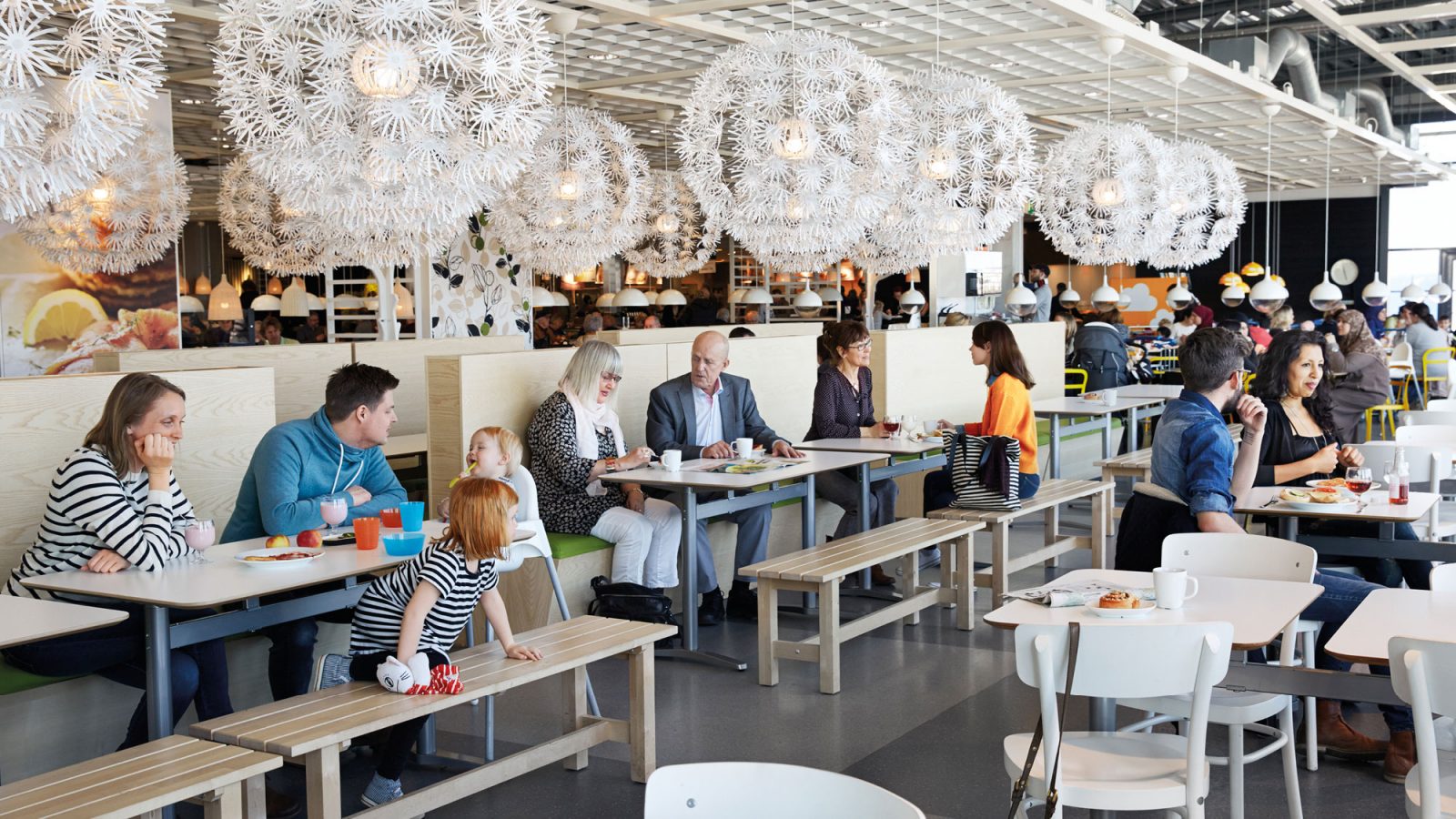 Familias con niños y parejas comen bajo grandes lámparas blancas en un amplio restaurante IKEA actual con muebles de madera.