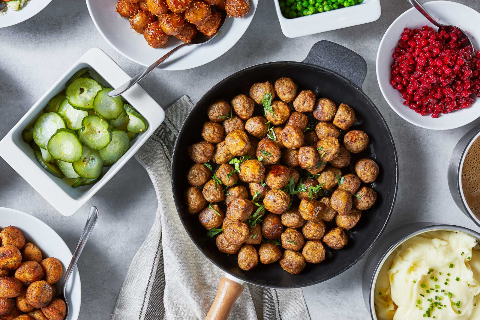 Tisch mit weißem Tuch, gefüllt mit Tellern und Töpfen bestehend aus Fleischbällchen, Preiselbeeren, Kartoffelpüree, Gurken und Soße.