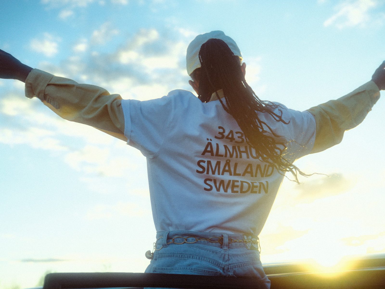 Frau, die an Autotür lehnt, in Jeans und T-Shirt mit schwarzem Aufdruck 343 36, ÄLMHULT SMÅLAND SWEDEN.