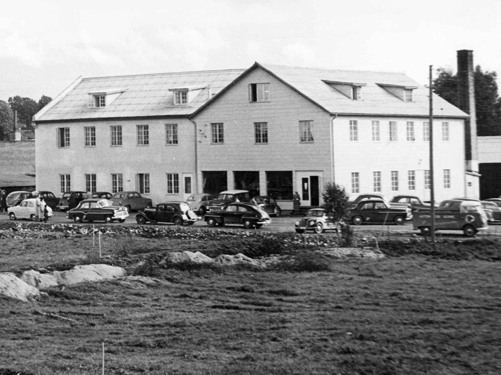 Großes weißes zweistöckiges Gebäude. Parkplatz mit Autos der 1950er. Im Vordergrund abgesteckte Baustelle.