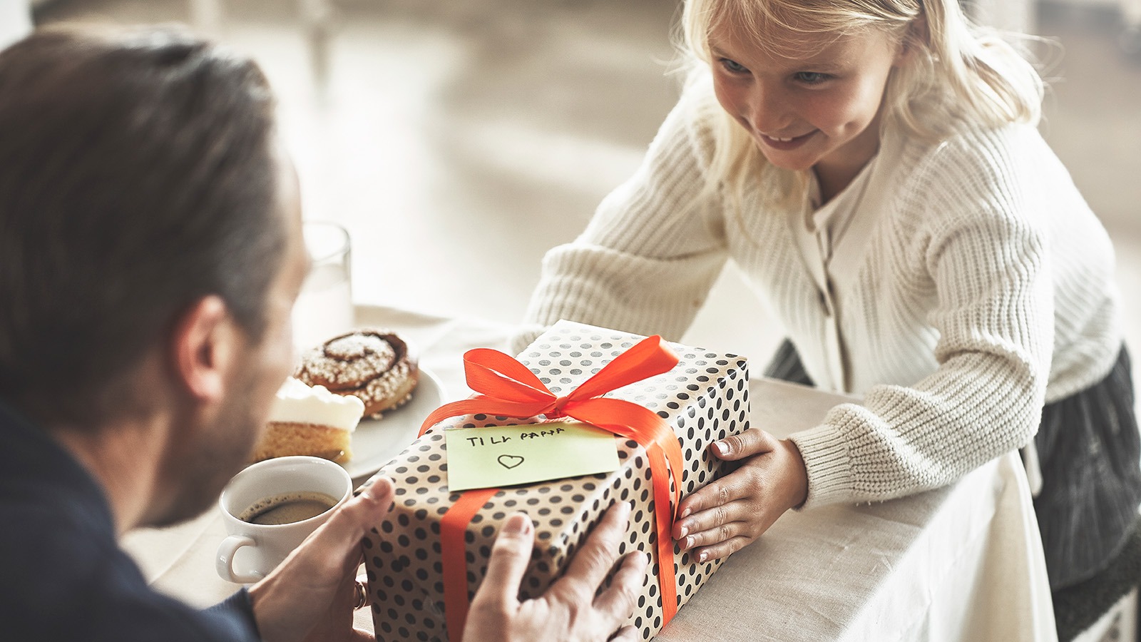 Litet barn räcker över ett inslaget prickigt paket med rött band till sin pappa.