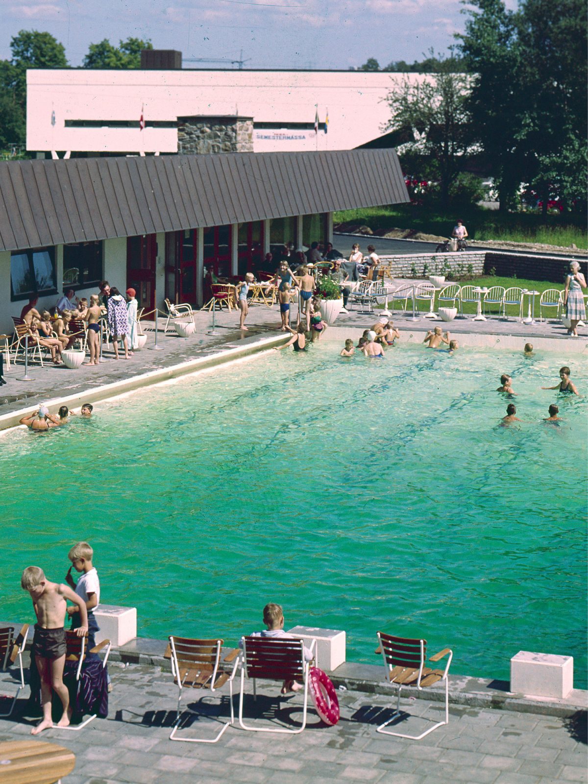 Large pool full of bathing children next to sunbeds and motel building, Motell IKEA. 1950s cars in background.
