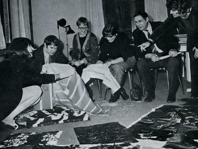 En grupp glada människor klädda i 1960-talsstil tittar på randiga tyger.