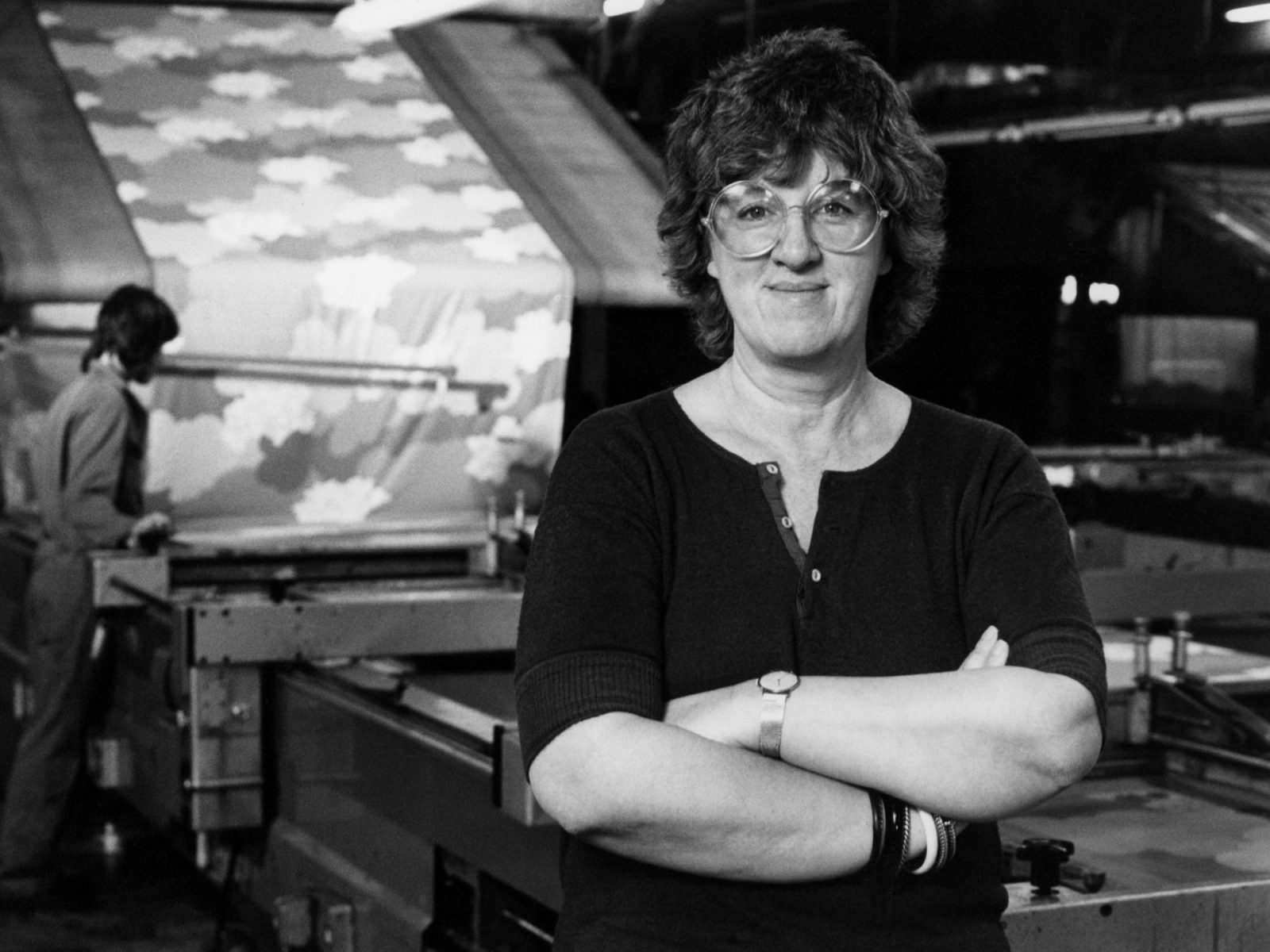 Kvinna med mörkt hår och stora glasögon i 1970-talsstil, Inez Svensson, står framför en textiltryckmaskin.