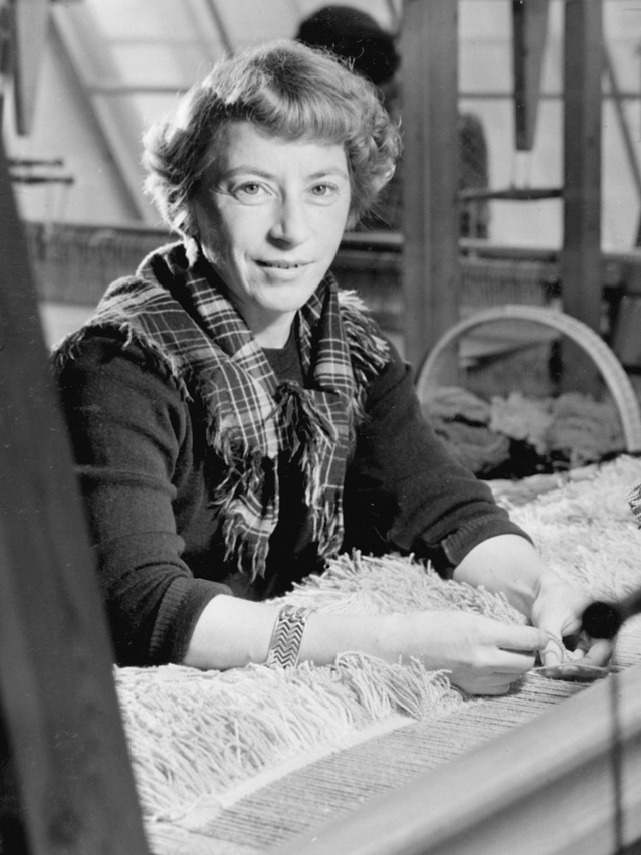 Viola Gråsten, en blond kvinna i 30-årsåldern, sitter vid en vävstol klädd i en tröja, rutig sjal runt halsen.